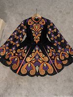 Full front view of the black Irish dance dress with colorful Celtic designs in orange, purple, and gold, laid flat showing long sleeves and flared skirt with scalloped hem edges.