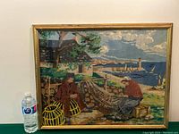 Full view of the framed needlepoint artwork in situ next to a water bottle for scale, showing the coastal scene with fishermen and lighthouse.