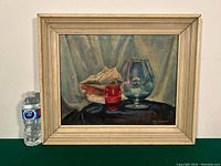 Framed still life oil on board painting featuring a conch shell, red glass, and large blue goblet with signature visible.