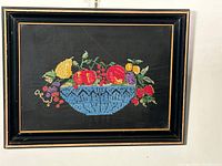 Full view of framed needlepoint artwork showing a colorful fruit bowl against a black fabric background, enclosed in a black and gold frame.