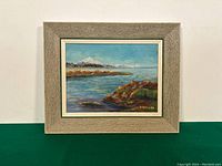 Framed oil painting of a coastal landscape showing Mount Baker with clear blue sky, water, and rocky shoreline. Signature M. Willam at bottom right.