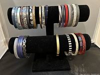 Two rows of 28 assorted bangle bracelets on a black velvet display stand, showing variety of colors and designs including metallics, patterns, and text.