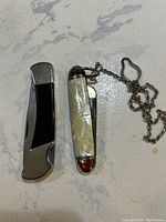 Two closed pocket knives, one with a black and silver handle and one pearlized with a chain.