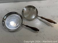 Two silver plated pans with wooden handles, placed side by side on a light colored surface, showing the front and side views.