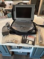 Open Apple PowerBook 160 laptop with keyboard, trackball, and screen visible. Power cord, software disks, floppy disks, manual, and carrying case on countertop.