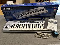 Photo of Edirol PCR-50 MIDI keyboard controller on the floor in front of its retail box, with power cord and manual placed beside it.