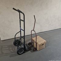 Two dollies side by side on concrete floor: large black dolly standing upright and small red dolly with a cardboard box on it.