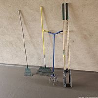 Four gardening tools against a wall: two leaf rakes, a garden claw, and a hole digger with long wooden handles.