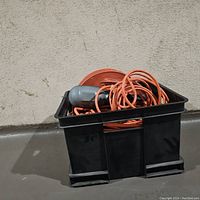Black plastic crate filled with several orange extension cords and one work light with attached cord.
