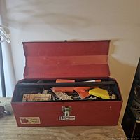 Front angle photo of the red metal toolbox open showing interior filled with various tools including drill bits, screwdriver handles, small saw, and hardware.