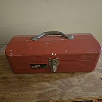 Closed red metal toolbox with silver latch and handle, showing wear and scratches on surface.