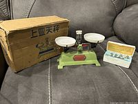 Mini balance scale with original box and balance weights in plastic case shown on a gray fabric couch.