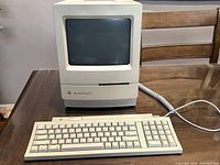 Full view of vintage Macintosh Classic II computer with keyboard on wooden table, showing front and side profile.