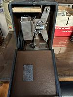 Vintage 8mm film projector inside a brown carrying case showing the metal construction and attached power cord.
