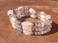 Close up photo of a triple strand bracelet composed of art glass beads with pink spots, clear faceted beads, and dark magnetic beads, laid on a wooden surface.