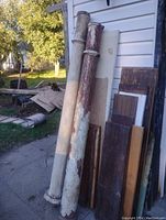 Two large antique wooden porch columns leaning against exterior siding next to stacked wooden panels. One column white, the other red.