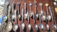 Top view of 13 silver plated souvenir spoons lined up showing colorful enamel crests and badges on handles.