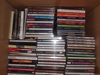 Overhead view of a box filled with stacked music CDs, showing variety in album artwork and case condition, primarily sealed with a few opened.