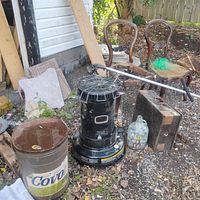 Photo showing kerosene heater, rusty metal pail, glass jug, Ryobi weed whipper, and two wooden chairs along with lumber and tiles in outdoor setting