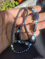 Close-up image showing varied shapes of polished hematite beads including cylindrical, teardrop, and faceted rectangular shapes, interspersed with bright blue round beads.