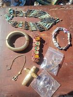 Photo showing the assortment of vintage costume jewelry laid out on a wooden surface including a turquoise and brass beaded necklace, metal bangle, two bracelets, necklaces with religious pendants, and earrings in plastic bags.