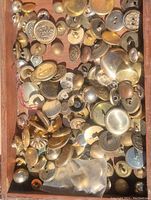 Close-up image showing numerous old metal buttons in brass and silver tones with various designs including military insignia and textured faces inside wooden box.