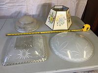 Four antique lamp shades displayed on a table with a measuring tape for scale. Three are glass with distinct patterns and one is a decorative hexagonal paper shade.