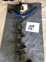 Full view of the gas powered earth auger with blue frame, mounted engine, auger bit attached, and instruction manual on a gray mat.