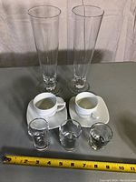 Front view of two tall glasses, two espresso cups on saucers and three shot glasses with measuring tape for scale