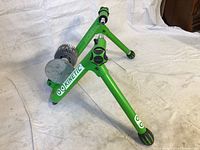 Oblique front-side view of the green Kinetic bike trainer, showing the resistance unit and the leg structure with rubber feet on a white cloth background.