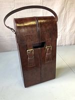 Brown leather wine carrying case with a single shoulder strap, two buckle closures at front, standing upright on white surface.