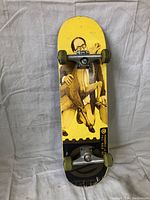 Front view of skateboard showing yellow deck with artistic graphic and green wheels mounted on metal trucks.