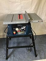 Front view of Ryobi 10 inch table saw, showing blue base, control panel with yellow/red switches, and black metal stand.