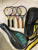 Four tennis rackets with two racket covers visible. The rackets are arranged with handles toward the bottom, showing a variety of colors and grip styles.