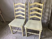 Two white wooden kitchen chairs side by side, showing ladder-back design and square seats.
