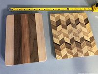 Two wooden cutting boards side by side with measuring tape showing about 11 inches width each. One board striped vertically with multiple wood tones, the other with a geometric chevron pattern.
