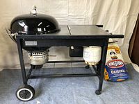 Full front view of the Weber charcoal grill on a black metal frame with wheels, gas ignition system beneath side compartment, and one full bag of Kingsford charcoal next to grill.