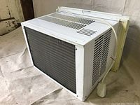 White window air conditioner unit viewed from the front and side, showing ventilation grilles and mounting hardware.