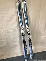 Full view of the two paired women's downhill skis standing upright showing white and blue tops with integrated bindings.