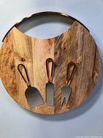 Top view of round wooden cheese board with natural grain and brass rim, showing three cheese tools attached with brass handles.