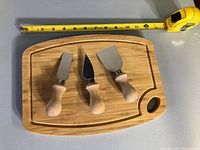 Rectangular wooden cheese board with three cheese knives displayed from above, tape measure showing approximately 12 inches long.