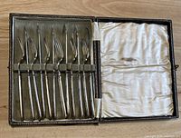 Opened original case showing six silver plated forks arranged in padded slots on green velvet lining, interior lid lined with white satin.