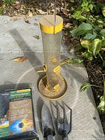 Photo of yellow and clear tube bird feeder with feeding ports and perches, and two hand garden tools with black metal tines placed on a concrete surface near plants.