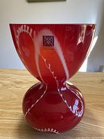 Front view of the red glass vase showing its hourglass shape with bulbous base and flared upper mouth, with glossy surface and some white wavy decorative lines.