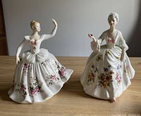 Two Royal Doulton porcelain figurines ‘Shirley’ and ‘Diana’ shown side by side, featuring detailed floral decorations and pink bows on their dresses.