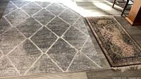 Photo showing both rugs on wood floor, the larger gray diamond rug on left and smaller eggplant rug on right.