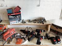 Photo of power tools on wooden shelf including Black & Decker belt sander, several cordless drills labeled Drill Master with batteries, chargers, and grinding attachments above shelf.