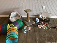 Overall view of the lot showing cat bed, tunnel, scratching post, feeding bowls, toys, and collar arranged on a wooden floor against a wall.