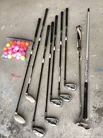 All golf clubs laid out on ground showing shafts and club heads along with a plastic bag of colorful golf balls.