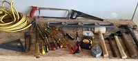 Full view of all hand tools including screwdrivers, hammers, mallet, saw, level, and extension cord on wooden surface against white background
